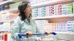 Mulher empurra carrinho de supermercado em frente a uma gôndola com diferentes marcas de creme dental