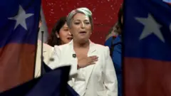Jeannette Jara, candidata presidencial en Chile, durante un acto electoral, se coloca la mano en el pecho, rodeada de banderas chilenas