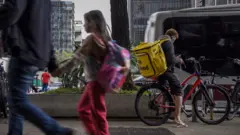 Homem e uma menina passando em movimento no primeiro plano; em segundo plano, com nitidez, um entregador está em uma bicicleta vermelha, usando uma bag amarela da 99, olhando para o celular
