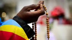 A hand holds a rosary. The edge of a Democratic Republic of Congo flag is draped over the person's arm.