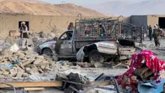 Un vehículo gravemente dañado, con luces de colores en los laterales, está rodeado por los escombros de un edificio destruido. La gente lo rodea inspeccionando la zona. Se ven montañas a lo lejos.