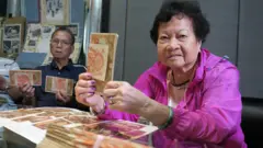 九龍深水埗一公寓樓住客會所內，身穿鮮粉紅色外套的香港索償協會前主席吳溢興遺孀李慧玲（右）舉起一疊紅色的100圓二戰日本軍票（BBC中文圖片）