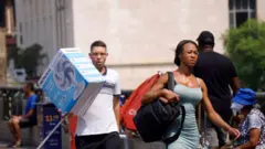 Pessoas andam nas ruas de São Paulo durante onda de calor. Uma mulher caminha de vestido de verão com corpo e rosto visivelmente suados, enquanto um homem pouco atrás dela carrega um ventilador numa caixa