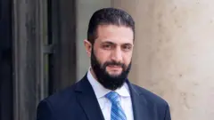 الشرع بلحية سوداء، مرتدياً بدلة رسمية كحلية اللون وقميص بلون أبيض وربطة عنق زرقاء مخططة، في قصر الإليزيه، يظهر خلفه حائط بلون سكري.