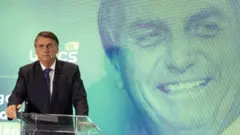 Bolsonaro com feição séria falando no microfone, com foto dele sorrindo atrás em painel