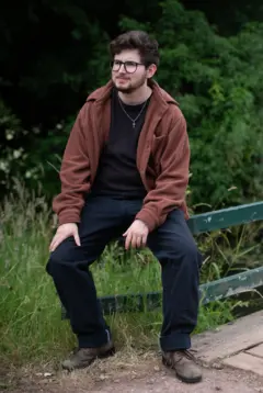 Homem vestido com casaco marrom e calças pretas, sentado ao lado de um grande campo verde, com arbustos ao fundo. Ele está sentado sobre um portão de madeira verde.