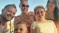 Foto de férias em família com um homem, uma mulher, duas meninas e um menino pequeno. Todos estão sorrindo.