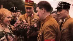Uma mulher de cabelos loiros trançados olha com admiração para Hitler enquanto ele fala com ela, cercado por homens em uniformes militares.