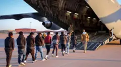 uçağa binmek için sıralanmış, elleri önden kelepçeli göçmenler