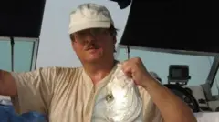 Um homem de bigode, usando óculos e um chapéu branco, segura um peixe pela cabeça