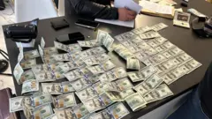На столі розкладено велику кількість купюр по 100 доларів США, частина складена рівними рядами, інша — хаотично. Поруч видно документи, телефони, гаманець і пристрій для перевірки банкнот, а одна людина тримає аркуші паперу.