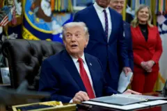 O presidente Donald Trump olha para o lado enquanto está sentado à sua mesa no Salão Oval, rodeado de pessoas.