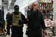 OMONchi va yuzini qo'li bilan yopgan ko'rinishidan rus millatiga mansub bo'lmagan kishi magazin ichida