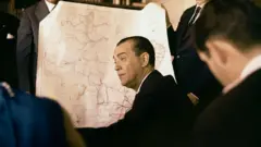 Um homem de terno, Juscelino Kubitschek, aparece sentado diante de um grande mapa, enquanto outras pessoas o observam ao redor.