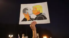 Um cartaz de protesto na Alemanha mostra ilustração de Putin e Trump se beijando