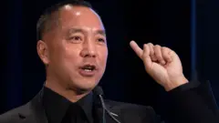 郭文貴在紐約一場新聞發佈會上發言，他身穿黑色西裝，舉起左手食指。（20/11/2018）