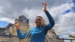 Yudi en una terraza con los brazos en alto y la batuta en la mano derecha moviendo sus largas trenzas blancas. Al fondo la fachada de un edificio señorial de Londres.