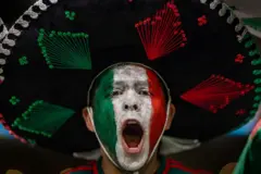 Un hombre con la boca abierta, seguramente está gritando o cantando. Tiene la cara pintada con los colores verde, blanco y rojo, que conforman la bandera de México. Lleva puesto un sombrero de mariachi.