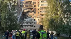 Пострадавшее жилое здание в Киеве была разрушена целая секция, лестница пожарных двигателей ведет к верхним этажам, на переднем плане являются сотрудники спасательных услуг