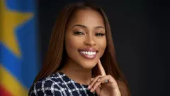 Grâce Kutino yambaye umwenda w'umukara n'umweru arimo guseka, inyuma ye hari ibendera rya DR Congo