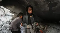 Žena u sivoj odeći sa crnom maramom na glavi stoji ispod ruševine. Iza nje dečak