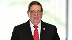 El ministro Bruno Rodríguez lleva puestos unos lentes y viste un traje oscuro, una camisa blanca y una corbata roja