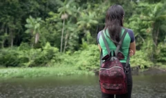 Meninapoker supremacostas com mochila e uniforme escolar observa o rio, com floresta ao fundo. 