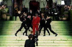 Grupo de personas baila y actúa en una escalinata verde durante un evento de gala; una persona en el centro viste traje rojo, rodeada de bailarines de negro, con músicos y cámaras al fondo.