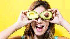 Happy young woman holding avocado halves - bright yellow background