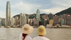 Eric y Emily de espaldas con sombreros sentados junto a un río. Al otro lado del río se ven edificios y rascacielos.