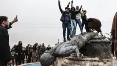 Rakka'nın Tabka ilçesinde yönetimin Suriye ordusuna geçmesini kutlayanlar, SDG tarafından konulan bir heykeli devirdi. Fotoğrafta devrilmiş savaşçı heykelinin üzerine çıkan ve zafer işareti yapan bir grup görülüyor. 