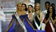 ブロンドの髪のミス・ユニバースの優勝者が、王冠と「ミス・ユニバース」と書かれたサッシュ、深紫のガウンを身にまとい、出場者たちから拍手喝采を浴びている