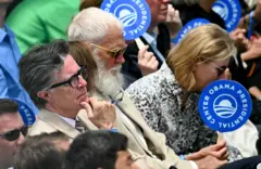 Stephen Colbert e David Letterman acompanham o evento cercados por outras pessoas. Algumas delas seguram cartazes circulares do Obama Presidential Center