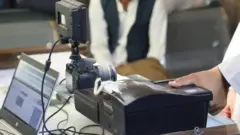 یک دستگاه الکترونیک ثبت انگشت که شست یک مرد روی آن قرار دارد، یک کمره و یک لب تاپ روی میز قرار دارد.