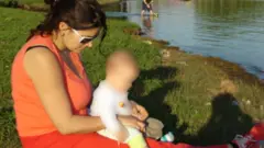 Raquel sits on grass near a body of water with her baby son, who is blurred, sat on her lap. She is wearing an orange t-shirt and trousers and white sunglasses. Her dark brown hair is half tied up at the back.