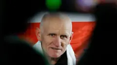 Крупным планом лицо Алеся Беляцкого на фоне бело-красно-белого флага белорусской оппозиции. При этом фотография обрамлена чем-то темным, снятым с очень близкого расстояния