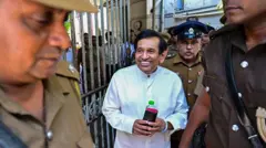 රාජිත සේනාරත්න අධිකරණයේ සිට රැගෙන යමින්. බන්ධනාගාර නිලධාරීන් පිරිසක් ඔහු වටා සිටිති