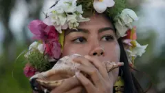 Una mujer de Guam, con una corona de flores tropicales sopla la concha de un caracol marino