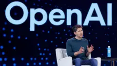 Sam Altman, CEO da OpenAI, sentado em uma poltrona branca, de camisa azul de mangas compridas e calça jeans, gesticulando com as mãos. Ao fundo, uma tela exibe o logotipo da OpenAI.
