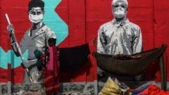 Grafite mostra dois trabalhadores de máscara; na frente da pintura, duas pessoas com mantimento aparentemente morando na rua
