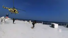 Un hélicoptère survole le pont d'un grand navire tandis que des hommes armés courent vers l'avant.