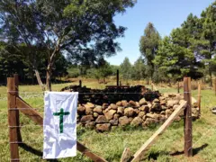 Imagem de um pano branco com desenho de uma cruz pendurado em uma cerca no campo