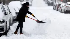 A man rugged up in heavy winter clothing uses a tool to clear snow packed into a shovel he is using to clear part of a road near a line of parked cars, all of which have snow over their roofs and windscreens.