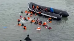 People wearing orange life jackets swim towards a black dinghy