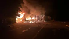 Уночі на дорозі палає зруйнована будівля або споруда. Яскраве полум'я охопило конструкцію, над нею піднімаються густі клуби диму. На асфальті перед пожежою видно уламки та дорожню розмітку зі стрілкою, що вказує напрямок руху.