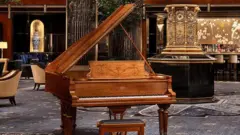 Un piano de cola de madera marrón en el centro, detrás un antiguo reloj dorado y butacas de madera con tapizado color crema.