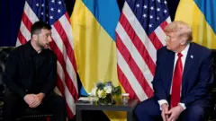 Зеленский что-то говорит Трампу. Они сидят у маленького столика с цветами и на фоне украинского и американского флагов