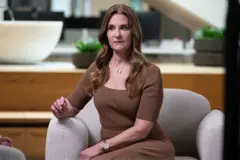 Melinda Gates é uma mulher branca, com cabelo longo e da cor castanho claro. Ela está usando um vestido marrom e sentada em uma poltrona cinza, durante uma entrevista.