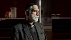 کشیش یعقوب کاسلیا در لباس سیاه کشیشی و یک کلاه سیاه مغموم و خشمگین به سمتی نگاه می‌کند