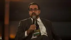 Christopher Garman sentado e falando ao microfone durante um evento.